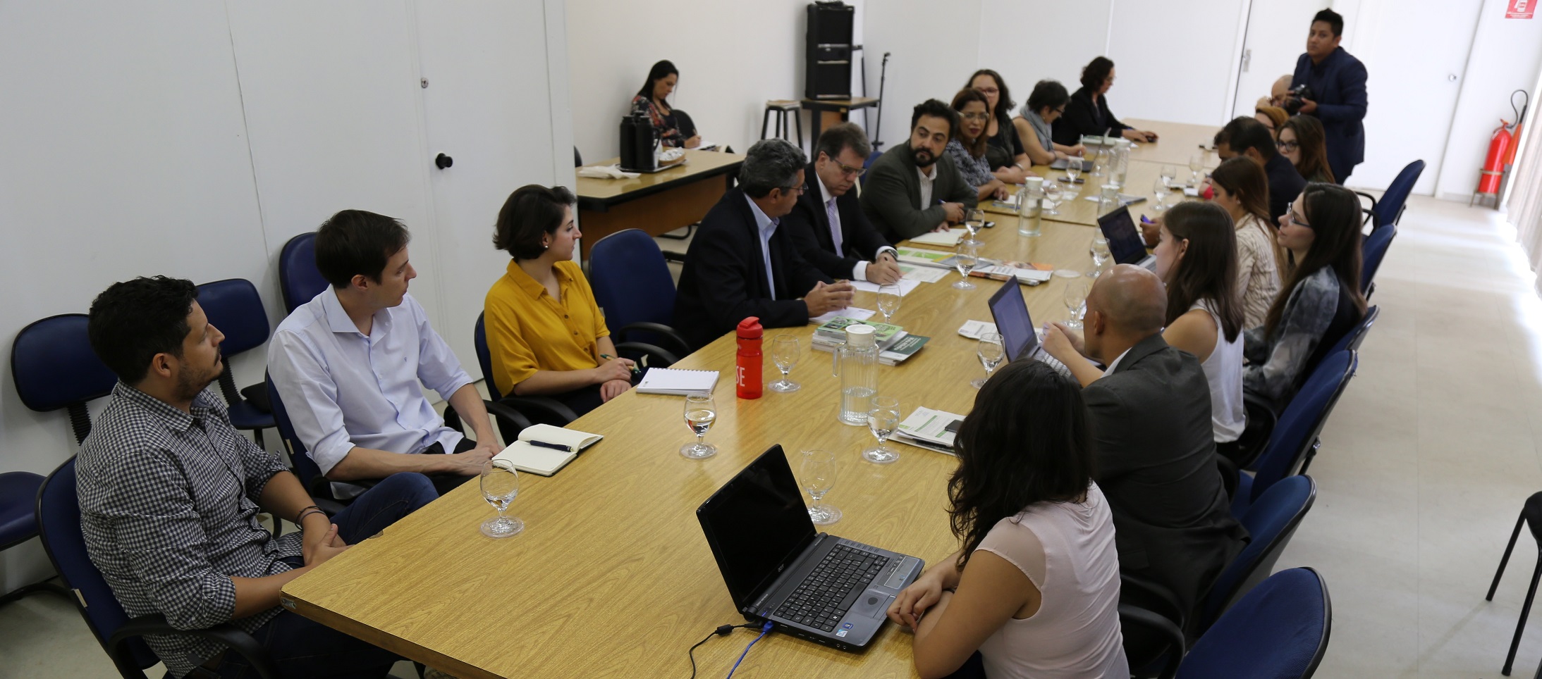 Participantes da reunião (homens e mulheres) sentados a mesa e conversando, alguns utilizam notebook.